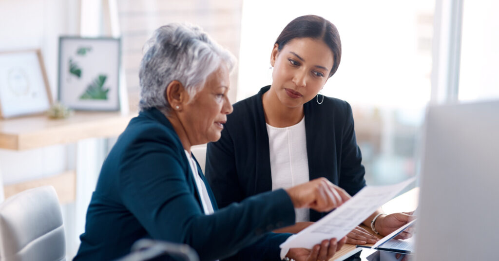 An older businesswoman shows a younger businesswoman a document